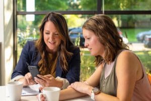 Two women at a table, deeply engaged with a cell phone, discussing Average Email Click Rates in E-commerce