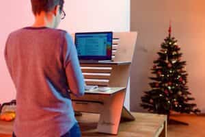 A man working on a laptop in front of a Christmas tree. The laptop is on a stand.